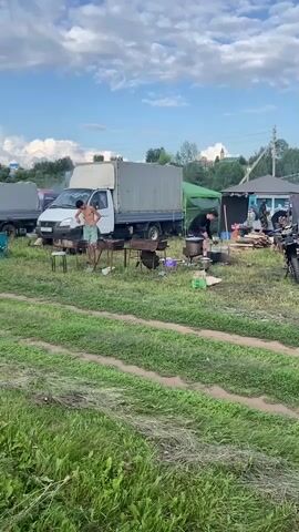 The video shows an outdoor gathering with several people and vehicles. In the foreground, there are rows of cut grass with a dirt path. In the background, there is a white truck with a grey tarp, a green tent, and a dark-colored gazebo. Several people are visible around these structures. Some are standing near grills, possibly cooking, while others are interacting or moving around the area. There are also some motorcycles and a car present. The environment appears to be a grassy field with trees and buildings in the distance, under a partly cloudy sky.