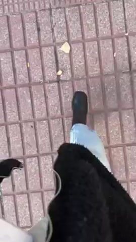 A first-person perspective video shows a person walking on a sidewalk. The sidewalk is made of different kinds of interlocking paving stones. The person is wearing blue jeans, black shoes, and a black, fuzzy jacket. A silver chain hangs from the jacket. The sidewalk is mostly red and tan, with some leaves scattered about.