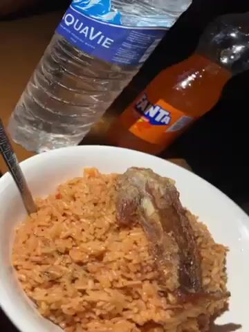 A close-up shot presents a bowl of jollof rice topped with a piece of meat, likely ribs. A metal spoon rests in the rice. Beside the bowl, there's a bottle of Aquavie water and a bottle of Fanta soda. The background is dark.