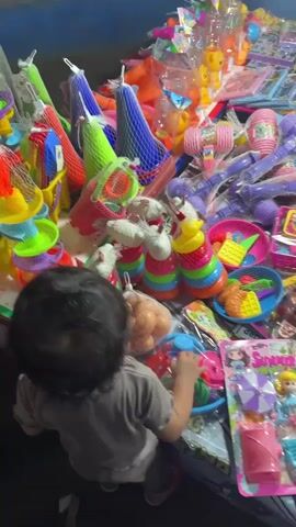 The video pans across a table laden with an assortment of colorful children's toys, presented in a market stall setting. A young child, seen from behind with dark hair, is positioned in the foreground, facing the table and reaching out towards the toys. The toys are diverse, including stacking rings, toy kitchenware, plastic food items, dolls, and various other playthings, many of which are packaged in plastic netting or cardboard. The lighting appears to be artificial, and the overall environment suggests an outdoor market or a busy indoor stall. The camera movement is fluid, capturing the sheer volume and variety of toys on display.