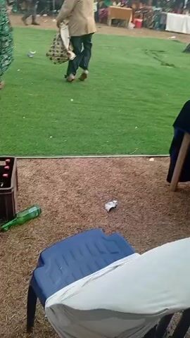 The video shows two men dancing on a green field in front of a crowd of people. The first man is wearing a camouflage outfit, and the second man is wearing a brown jacket and dark pants and carrying a bag. There are tents set up in the background. There are also cases of bottled drinks and an empty green bottle in the foreground. The man in the brown jacket takes off his jacket and continues to dance.