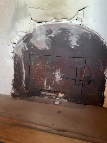 A close-up shot focuses on the rustic metal door of a wood-fired oven. The door is arched at the top, made of dark, weathered metal with visible scuff marks and scratches. A simple latch mechanism secures the door, and a small gap at the bottom allows a faint glow of embers or fire to be seen. The oven opening itself is built into a rough, textured wall, possibly made of stone and plaster, with a creamy white finish that shows signs of age and wear. The surrounding hearth appears to be made of concrete or a similar material, with a wooden element visible at the bottom of the frame. The lighting is dim, suggesting an indoor setting, and the overall impression is one of a well-used, traditional cooking appliance.