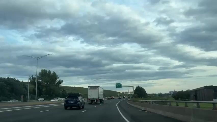 The video shows a view from a car driving on a highway. The sky is cloudy with gray and white clouds. There are several cars and trucks on the highway. A green sign indicating "EXIT 32 Jennings Road 1/2 MILE" is visible over the highway. To the left of the highway, there is a grassy hill with trees. To the right, there is a concrete barrier and some trees. The overall impression is of a typical drive on a cloudy day.