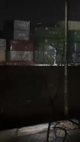 The video shows a night scene of stacked shipping containers. The containers are various colors including red, green, and beige. Some are labeled with shipping company names like "CHIYU" and "HANJIN SHIPPING".  A dark wall and bare branches are visible in the foreground.  A utility pole stands at the right. The ground appears to have patches of snow or ice.  The lighting is poor, but suggests streetlights or other sources are present.  The camera is stationary.