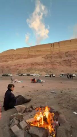 A video shows a group of people gathered outdoors, sitting around a campfire. In the foreground, a campfire burns brightly, surrounded by rocks. A young woman with dark hair, wearing a white sweater and light-colored scarf, sits on a wooden object next to a young man. The man is sitting on the same wooden object and is wearing a dark-colored jacket. They are facing the camera, but only the back of the man's head is visible. In the background, a large, layered rock formation rises, with people and vehicles scattered across the flat ground in front of it. The sky is blue with white clouds tinged with sunlight. The atmosphere appears relaxed and social.