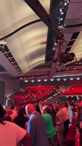 This video captures a vibrant event taking place inside a large, modern venue, likely a church or convention center, in Dallas, Texas. The scene is filled with people, both on a stage at the front and in the audience filling tiered seating that curves around the space.

On the stage, several individuals are visible, seemingly performing or speaking. One person is in the foreground of the stage, holding a microphone and gesturing. Behind them, another person is seen, and further back, a group of individuals are gathered. A large screen on the stage displays an image of a person holding a microphone. Another large screen to the right of the stage also shows a similar image. The stage is illuminated with dramatic lighting, including spotlights and colored lights.

The audience is densely packed, with many individuals facing the stage. The mood appears energetic and engaged, with some people in the audience raising their hands. The overall lighting in the venue is a mix of stage lighting and ambient light, creating a dynamic atmosphere. The architecture of the venue is modern, with a curved ceiling structure that adds to the spacious feel.

Visible text on the stage screen reads "HALLELUJAH". The time of day is not explicitly indicated, but the indoor setting and lighting suggest an evening or indoor event. There are no clear indications of weather. The location context of Dallas, USA, is provided.