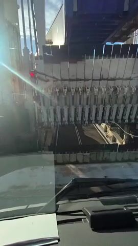 The video shows a view from inside a vehicle, looking out through the windshield at a large industrial machine. The machine appears to be a conveyor belt system with a series of vertical, parallel metal components that could be part of a sorting or processing mechanism. In the upper left of the frame, a red light is visible, possibly indicating a stop signal or an operational status. The overall environment seems to be outdoors, with sunlight casting shadows and creating lens flare on the windshield. The machine is likely automated, with no visible human operators.
