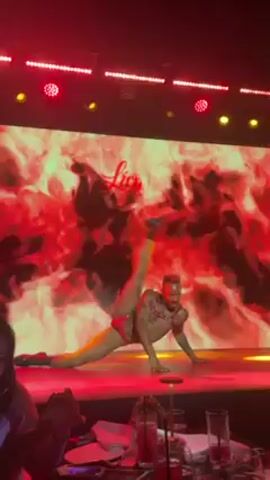 Here is a description of the video:

The video shows a dancer performing on a stage in what appears to be a nightclub or entertainment venue. The dancer, wearing minimal red attire and black boots, executes a series of gymnastic and dance movements, including splits, handstands, and leg extensions. The stage is illuminated by red lighting and features a large video screen displaying abstract imagery resembling fire or smoke, with the word "lio" overlaid. The audience is visible in the foreground, seated at tables with drinks. The overall atmosphere is lively and suggestive, typical of a nightlife setting.