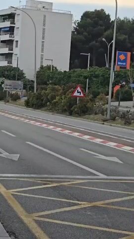 A black Jeep Compass travels along a paved road. It passes by the camera, which is situated at the roadside. A sign indicating the direction to Barcelona can be seen. In the background, there is a multi-story building and a gas station.