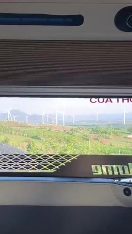 The video captures the view from inside a moving vehicle, likely a bus or van, looking out a large window at a scenic landscape. The scene outside features numerous white wind turbines spread across verdant, hilly terrain, with a body of water visible in the distance. Green foliage and trees dot the landscape under a bright, clear sky, indicating daytime.

Inside the vehicle, the interior appears modern and well-maintained, featuring cream-colored trim and a grey dashboard area. Above the window, a textured shade or panel is visible, along with air vents and two cup holders. Below the window, on a dashboard-like surface, there are several control buttons, some illuminated with blue light, and a small digital display. A person's hand briefly enters the frame to the left, seemingly adjusting something near the window.

Text is visible both directly and as a reflection. In red lettering above the window, "CUA THOAT HIEM" (Vietnamese for "Emergency Exit") is clearly readable. Below the window, reflected on the dashboard, partially visible and reversed text resembling "9mollido" or "Mobilido" can be seen. The vehicle is in motion, with the exterior scenery steadily passing by.