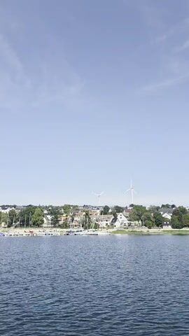 A wide shot captures a calm blue body of water in the foreground, with a treelined shore and a distant town visible in the background under a clear blue sky. The shore is dotted with various buildings and wind turbines. Numerous sailboats are moored along the shoreline. The video is short and shows no distinct actions or interactions.