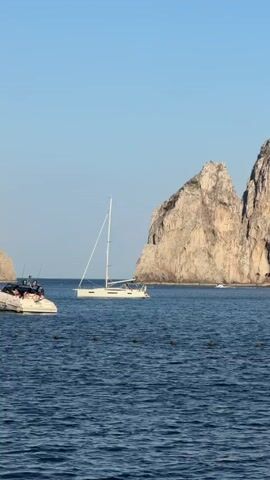 The video shows a seascape with two prominent boats and a striking rock formation in the background. A white sailboat with its mast raised is visible in the center of the frame. A small dinghy is attached to the sailboat. People can be seen on the sailboat. Another motorboat is seen to the left of the sailboat. In the background is a large, rocky land formation, possibly a cliff or island, split into two distinct peaks, that stands out against the clear blue sky. The sea is calm with gentle waves and there is green seaweed floating on the surface. The environment suggests a sunny, open-water scene.