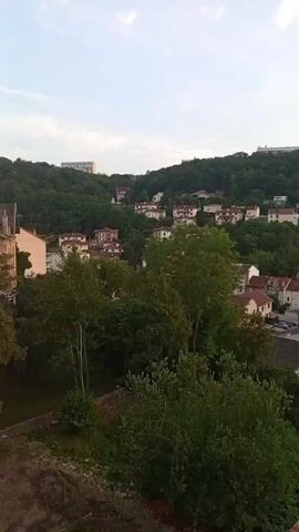 The video shows a panoramic view of a hillside town with houses scattered among trees. The sky is partly cloudy, with the sun beginning to set on the right side of the frame. The camera pans slowly from left to right, revealing more of the town and the surrounding landscape. The overall impression is one of a peaceful evening in a suburban area.