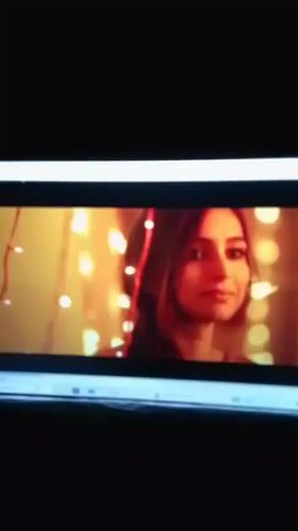 A video plays on a screen. The first frame shows a woman with shoulder-length brown hair and brown eyes, wearing a black top. She is looking to her right with a faint smile. The background is blurred with yellow and white lights. The next few frames show an audience in a dimly lit venue. A man in the foreground wears sunglasses and a grey jacket. A woman to his left wears an orange and yellow sari. The final frame shows a group of people gathered around a man in a suit, who is holding a microphone and speaking. The background is illuminated by fairy lights and some yellow spotlights.