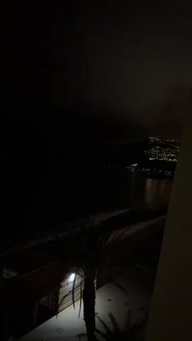 The video shows a dark scene at night, presumably taken from a high vantage point. A body of water, likely a sea or ocean, is visible, with the faint reflection of lights shimmering on the surface. In the distance, there's a hillside or landmass dotted with numerous lights, suggesting a town or city. The foreground features a beach and some vegetation, possibly palm trees, though details are obscured by the darkness. A building structure is partially visible on the right side of the frame.