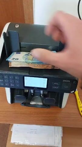 The video shows a money counting machine on a wooden shelf. The machine is counting a mix of 20 and 50 Euro banknotes. The display shows the count increasing as the machine processes the money. There is a yellow highlighter and some papers on the shelf next to the machine. The background includes a wooden door or wall.