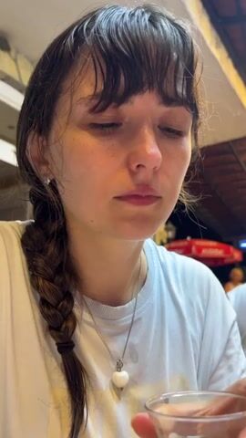A close-up shot shows a young woman with dark hair in a braid and bangs, drinking from a small, curved glass of dark liquid, possibly tea. Her eyes are wide and her head is tilted slightly as she sips. She is wearing a white t-shirt and a necklace with a white heart-shaped pendant. The background is blurred, but appears to be an indoor setting with warm lighting. The woman’s facial expression suggests contemplation or enjoyment of the drink.