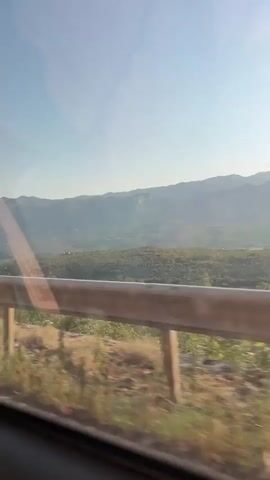 A view from a moving vehicle shows a mountainous landscape under a clear sky. In the foreground, a metal guardrail runs horizontally across the frame, partially obscuring the view. The guardrail is made of two parallel metal bars supported by vertical posts. Beyond the guardrail, a sloping terrain is covered with sparse vegetation and trees. In the distance, rolling hills and mountains recede into the haze, with the sky above being a pale blue. The motion blur indicates the vehicle is in transit.