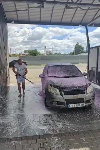 A bald man in a white t-shirt and white shorts is pressure washing a car. The car is covered in pink foam. The man is holding a pressure washer hose and spraying water on the car. The car is in a car wash bay with a metal roof and walls. There is foam and water on the ground. The sky is visible in the background with clouds.