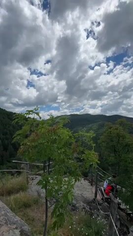 The video shows a panoramic view of a lush green mountainous landscape under a partly cloudy blue sky. In the foreground, there are trees and rocky outcrops, and a wooden railing with a rope barrier suggests a viewpoint or hiking trail. Two people, one wearing a red top, are seated on a wooden bench along the railing. The camera pans slowly, revealing more of the rolling hills and distant mountains. The overall impression is of a scenic and serene natural environment.