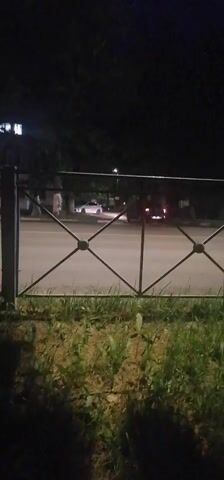 A white car turns right onto a street at night. A dark-colored car is parked on the side of the road, its brake lights are visible. A metal fence separates the camera from the street. The area is grassy and dark.