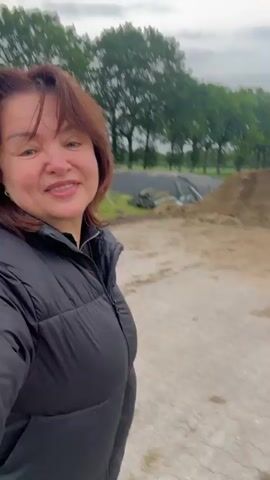 A woman with auburn hair, wearing a black puffer jacket, smiles directly at the camera. She is outdoors in what appears to be a rural or agricultural setting. Behind her, there is a large pile of dirt or hay, and in the background, trees and what looks like a covered silo or storage facility. The ground in front of her is a hard surface, possibly concrete or asphalt, with some damp patches. The lighting is overcast. The woman's smile is warm and friendly.