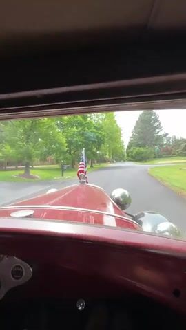 The video shows the interior view of a red vintage car moving down a quiet residential street lined with green lawns and trees. The car's dashboard is visible, featuring a steering wheel, gauges, and chrome details. An American flag is mounted on the front of the hood, visible through the windshield. The driver's hands are on the steering wheel, and the car appears to be moving at a slow pace. The sky is overcast, casting a soft light on the surroundings.