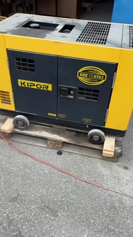 The video begins with a close-up of a yellow and black KIPOR KDE12STA3 diesel generator sitting on a wooden pallet with four small wheels. The generator has a black door with a silver handle and a ventilation grill. The camera then pans left to show a black forklift with its forks extended, positioned near the generator. The forklift is in front of a warehouse-like building with shelves stacked with boxes. The video continues to pan left, showing more of the forklift and the warehouse interior. The forklift's mast is visible, and there are various boxes and items on shelves in the background. The scene is outdoors, on a paved surface, with a red cable running from near the generator towards the left of the frame. The lighting suggests it is daytime.