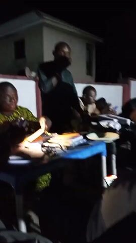A group of people are gathered around a blue plastic table outside at night. A man in a dark shirt is standing and speaking, gesturing with his hands. Some of the other people at the table are looking at their phones, while others have notebooks and papers in front of them. There are also plates with food on the table. A white building with a dark roof is visible in the background.