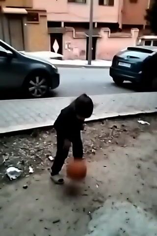 A young child dressed in dark clothing is dribbling an orange basketball in an outdoor setting. The child is positioned between a sidewalk and a dirt area with scattered debris. Two cars are parked on the street next to the sidewalk. Buildings are visible in the background.