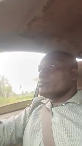 A man is driving a car. He is bald, wearing a light-colored collared shirt and a tie. The car's interior appears worn. Trees and vegetation are visible outside the car, blurred due to motion. The sky is overcast.