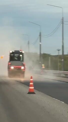 A road roller is compacting asphalt on a road. The roller is orange and black with flashing lights on top. It is kicking up a lot of dust. Several people in high-visibility vests are working on the road. A traffic cone is in the foreground. The sky is overcast and there are utility poles in the background. The video appears to be taken from a moving vehicle.