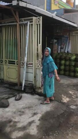 A woman in a patterned dress and headscarf is sitting outside a store. She is looking towards the left of the frame. Behind her, a woman in a black headscarf and brown top is standing. In front of the seated woman are several large blue water jugs. To the right of the jugs are stacks of green propane tanks. To the left of the seated woman are stacks of boxes and a person wearing a blue hijab is walking past the foreground. The ground is concrete and wet. The store appears to be selling bottled water and propane.
