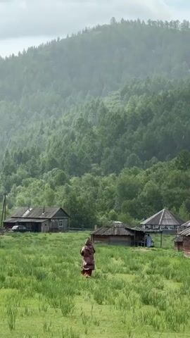 In a rural village setting, a person wearing a brown cloak stands in a grassy field. Several wooden houses with dark roofs are visible in the background. A lush, green forest covers the rolling hills behind the village. Light rain is falling, indicated by streaks in the air. Another person in a blue garment is partially visible near a structure on the right.
