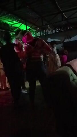 The video shows a group of people dancing at what appears to be an indoor club or outdoor patio with a roof. The lighting changes between green, blue, and red. Most of the people are dressed casually and moving to the music. A person in a pink hoody, red and white patterned shirt, and dark pants is dancing prominently in the center. The people in the scene are obscured by shadows and the video quality is low.