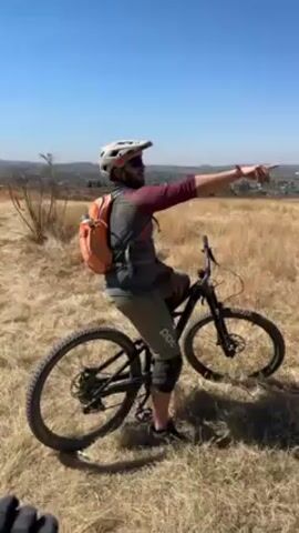A man on a mountain bike points toward a distant view of a snow-capped mountain, then the camera pans to the right, revealing a dry, grassy hillside. The man is wearing a helmet, sunglasses, and a backpack. The bike is black with disc brakes. The sky is clear and blue.