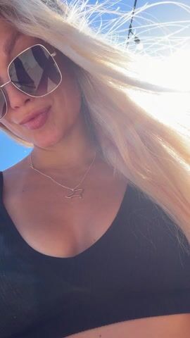 A woman with long blonde hair smiles into the camera while standing outdoors. She wears a black bikini top and large sunglasses. In the background, two men stand at a DJ setup, operating the equipment. The setting appears to be an elevated outdoor event with a view of mountains in the distance under a clear blue sky.