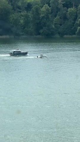 This is a short video clip captured from a high vantage point overlooking a body of water, likely a lake given the presence of a treed shoreline. The scene is set in Mikołajki, Poland.

The primary subjects are two boats navigating the water. The larger boat, a dark-colored motor yacht, is positioned on the left side of the frame and is moving to the right. It's creating a noticeable wake behind it, indicating it's in motion. To the right of the yacht, a smaller, lighter-colored boat, possibly a speedboat or a similar craft, is also moving in the same direction. This smaller boat is trailing the larger one and also generating a wake.

The setting is a wide expanse of water with a distinct green hue, suggesting it might be a lake or a calm river. The background is dominated by a dense, lush forest line that stretches across the horizon, with various shades of green indicating a variety of trees. The shoreline appears to be quite close to the boats, suggesting the video is taken from a moderately elevated position, possibly a balcony or a hill.

The weather appears to be overcast or hazy, as the light is diffused and there are no harsh shadows. The time of day is not explicitly indicated but the lighting suggests it could be daytime, possibly mid-morning or afternoon. There are no people clearly visible on either boat, but their presence is implied by the operation of the vessels.

There is no visible text within the video frame. The overall mood is serene and suggests a peaceful day on the water, with the boats enjoying the natural beauty of the Polish landscape.