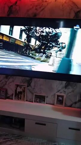 A dark flat-screen television is displaying an action scene of a giant robot in a city, possibly from the movie Transformers. The TV is mounted on a white marble-patterned wall, which is illuminated by red and green LED strip lights along the top. Below the television, a white entertainment console with drawers is visible. Several framed photographs are displayed on top of the console. The floor is covered with a patterned rug.
