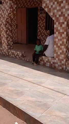 An adult male and a young boy are seated on a tiled porch in front of a building during daylight hours. The adult, wearing a white shirt, is positioned on the right, facing away from the camera. The boy, dressed in a green shirt and dark trousers, sits to his left, facing forward. They appear to be resting or waiting.

The building features a distinctive brown and beige checkerboard tile pattern covering its exterior walls, pillars, and the porch. A wooden door is visible in the center of the building's entrance. The ground in front of the porch is also tiled with larger, light-colored square tiles. Strong shadows suggest bright, sunny weather. The setting is outdoors, likely in an urban or semi-urban area. No discernible text is clearly visible on the building or in the scene.