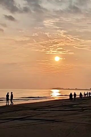 The video depicts a beach scene at sunset.  The sun is visible in the sky, partially obscured by clouds.  Several people are present on the beach.  Two individuals stand at the water's edge, appearing to look out at the ocean.  A group of people is walking along the shoreline in the distance.  The beach itself is sandy, and there are tire tracks visible in the sand. The ocean reflects the sunlight.