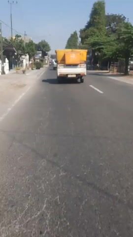 A white pickup truck with a yellow cargo box is driving on a paved road. The truck has a license plate that appears to be yellow with black lettering. The road has white lane markings. There are trees and buildings on the side of the road. The sky is blue and clear. The video is shot from a moving vehicle behind the pickup truck.