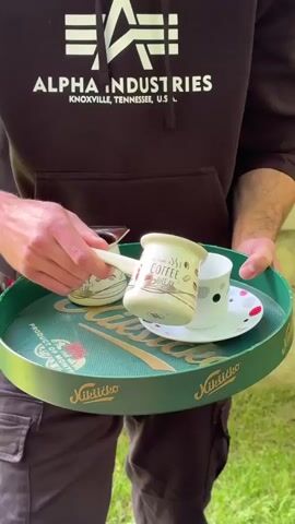 A young man wearing a black hoodie with an "Alpha Industries" logo and black cargo pants is standing outdoors and holding a green tray. On the tray, there are two small white mugs with a "Coffee Break" design and a white teacup with a colorful polka-dot saucer. The man is pouring dark liquid, presumably coffee, from one of the mugs into the teacup. The tray has some text and graphics on it, including "Nikšićko" written on the side. The background shows greenery and a building. The camera is focused on the man and the tray.