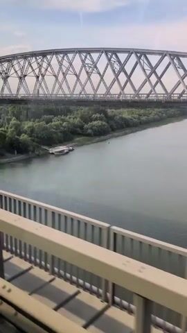 This video shows a wide river with a truss bridge overhead. The camera is positioned on a lower bridge or walkway, looking across the river. The river is calm, with a greenish-gray hue. On the far bank, a dense line of trees runs along the water's edge. Two boats are docked near the bank, one larger than the other, appearing to be houseboats or tour boats. The truss bridge is a prominent structure, made of interconnected metal beams forming a geometric pattern. The sky above is partly cloudy with a bright, diffused light, suggesting daytime. The video appears to be shot from a moving vehicle, as indicated by a slight blur and the perspective shifting subtly.