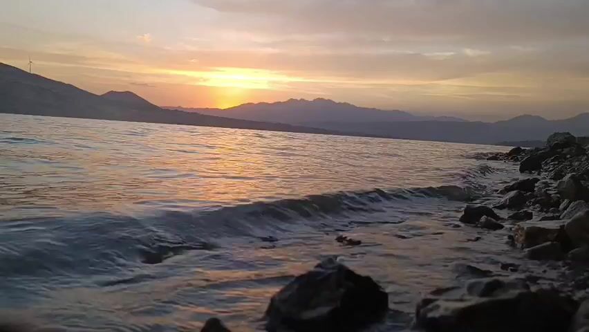 A low-angle shot of a body of water near a rocky shore during sunset. The sun is visible on the horizon, casting a warm glow on the sky and water. Mountains form a silhouette in the background. The water is gently lapping at the shore, creating small waves. There are a few wind turbines visible on a distant hill. The overall scene depicts a tranquil natural landscape at dusk.