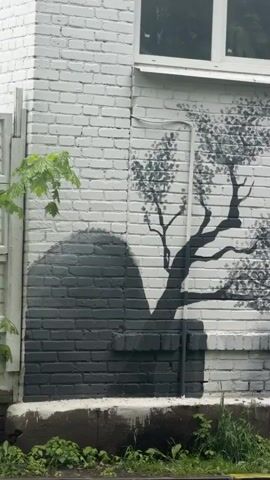 The video showcases a mural painted on a white brick wall. The mural depicts a black and white landscape featuring trees, a pagoda, a bridge, and a rocky hill. The word "KARATE" is written in black above the hill. A white drainpipe runs vertically along the wall, partially obscuring the painted tree. Green vegetation grows along the base of the wall. A white-framed window is visible above the mural.