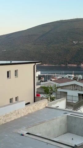 This media file captures a serene panoramic view of a coastal area, likely a harbor or bay, with a lush, tree-covered mountain range dominating the background. The setting appears to be a developed residential or resort area, with several modern buildings in various stages of completion.

In the foreground, a partially constructed rooftop area is visible, featuring a concrete pool basin lined with what appears to be marble or stone tiles. Building materials, possibly tiles, are stacked neatly on the rooftop. Adjacent to this, several multi-story residential buildings are present, characterized by clean, contemporary architectural designs with white facades and stone accents. Some of these buildings have balconies and large windows, suggesting they are intended for habitation.

Beyond the immediate buildings, the scene opens up to a body of water filled with numerous sailboats and yachts moored in a harbor. Cranes, indicative of ongoing construction or development, are visible in the distance along the waterfront and on adjacent land, suggesting this is a dynamic area undergoing expansion.

The time of day appears to be late afternoon or early evening, as indicated by the warm, soft lighting and the gentle illumination of the landscape. The sky is a clear, pale blue, suggesting fair weather. The overall impression is one of tranquility and natural beauty, juxtaposed with ongoing development and human activity. No people are visible in the frame, and there is no discernible text in the media. The location cues, such as the style of architecture and the mountainous coastal landscape, along with the provided location context of Đenovići, Montenegro, align with this visual assessment.