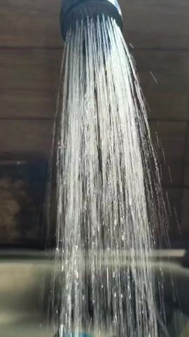 This is a close-up, upward-facing shot of a showerhead with water spraying out. The water streams are thin and separated, creating a mist-like effect as they fall. The background is blurred but appears to be a tiled surface, possibly a shower wall or bathroom fixture. The lighting is bright, highlighting the water droplets.