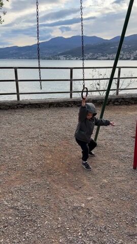 The video shows a young child, probably a toddler, playing on a playground next to a body of water. The child is wearing a grey hooded jacket, black pants, and grey shoes. The child is holding onto a metal ring hanging from a chain. The playground surface is gravel. A metal railing and some sparse vegetation separate the playground from the water. In the background are mountains and a cloudy sky. The child attempts to swing but is too small and appears to lose their balance slightly.
