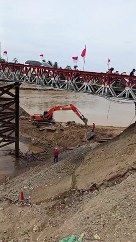 The scene is dominated by an orange excavator operating on a sandy bank next to a muddy river. The excavator is actively digging and moving material. Two individuals in red shirts and hard hats are visible near the excavator, potentially directing or observing the operation. Above them, a steel truss bridge, painted with alternating red and white sections, spans the river. Several vehicles, including a white SUV, are crossing the bridge, and people can be seen standing on it. Red flags are mounted along the length of the bridge. The sky is overcast, and the overall environment suggests an active construction or maintenance project occurring near the river. A brown, dirt hillside is in the foreground, adding to the industrial feel of the scene.